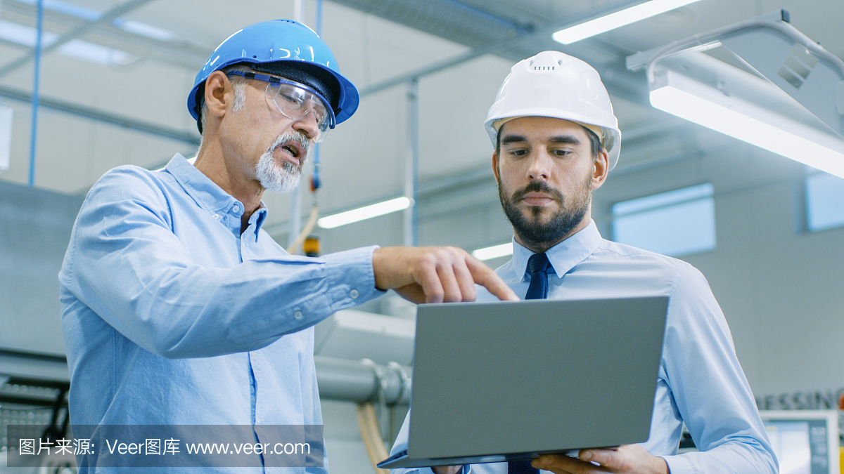 部門主管持有筆記本電腦并與總工程師討論產品細節。他們戴著安全帽在現代工廠工作。